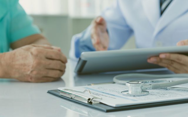 Photo of consultation, doctor holding an iPad and patient sitting on left with hands crossed. Photo of stethescope in the table on top of clipboard.